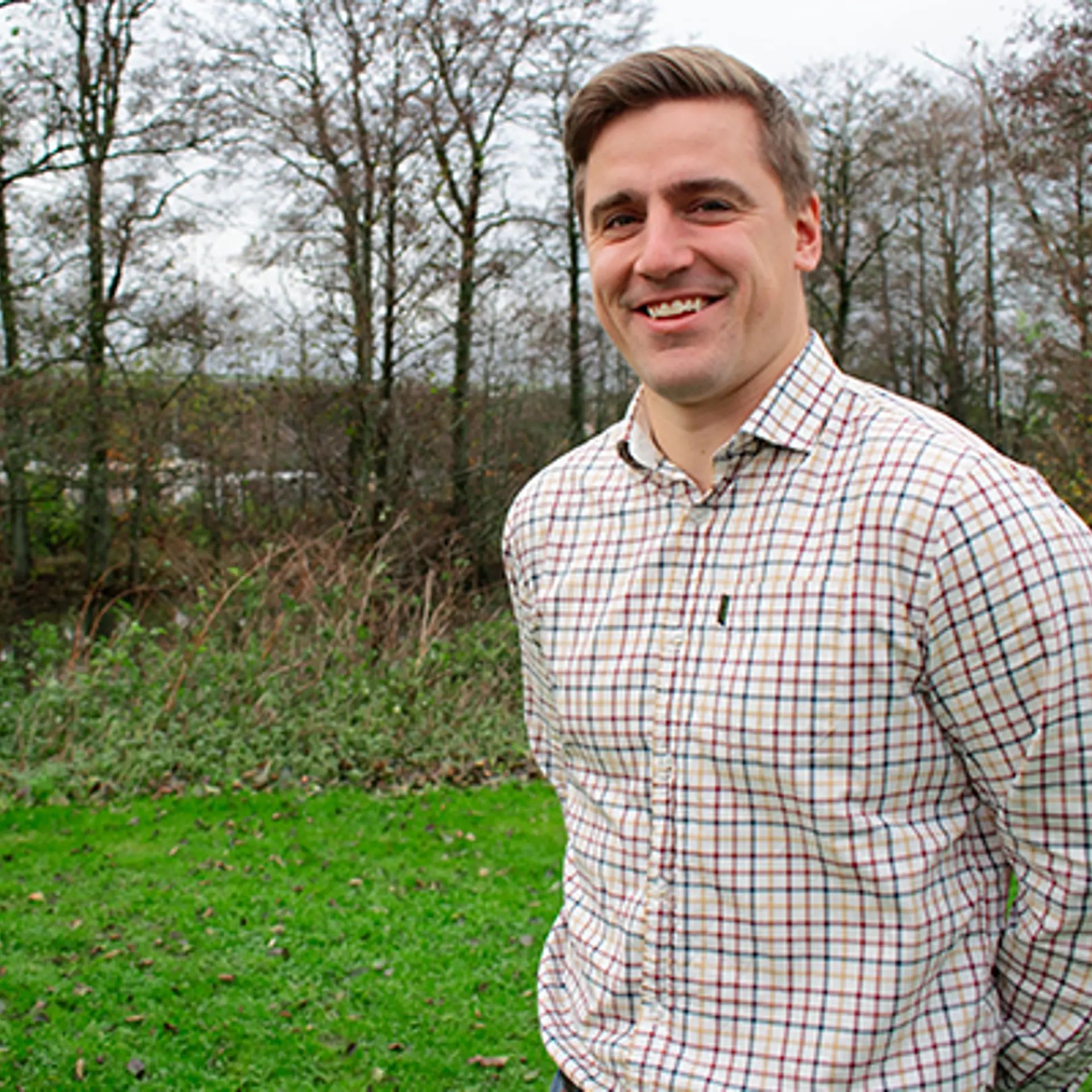 En yngre man i mörkt hår ler. Han bär en beige rutig skjorta. han befinner sig utomhus på hösten.