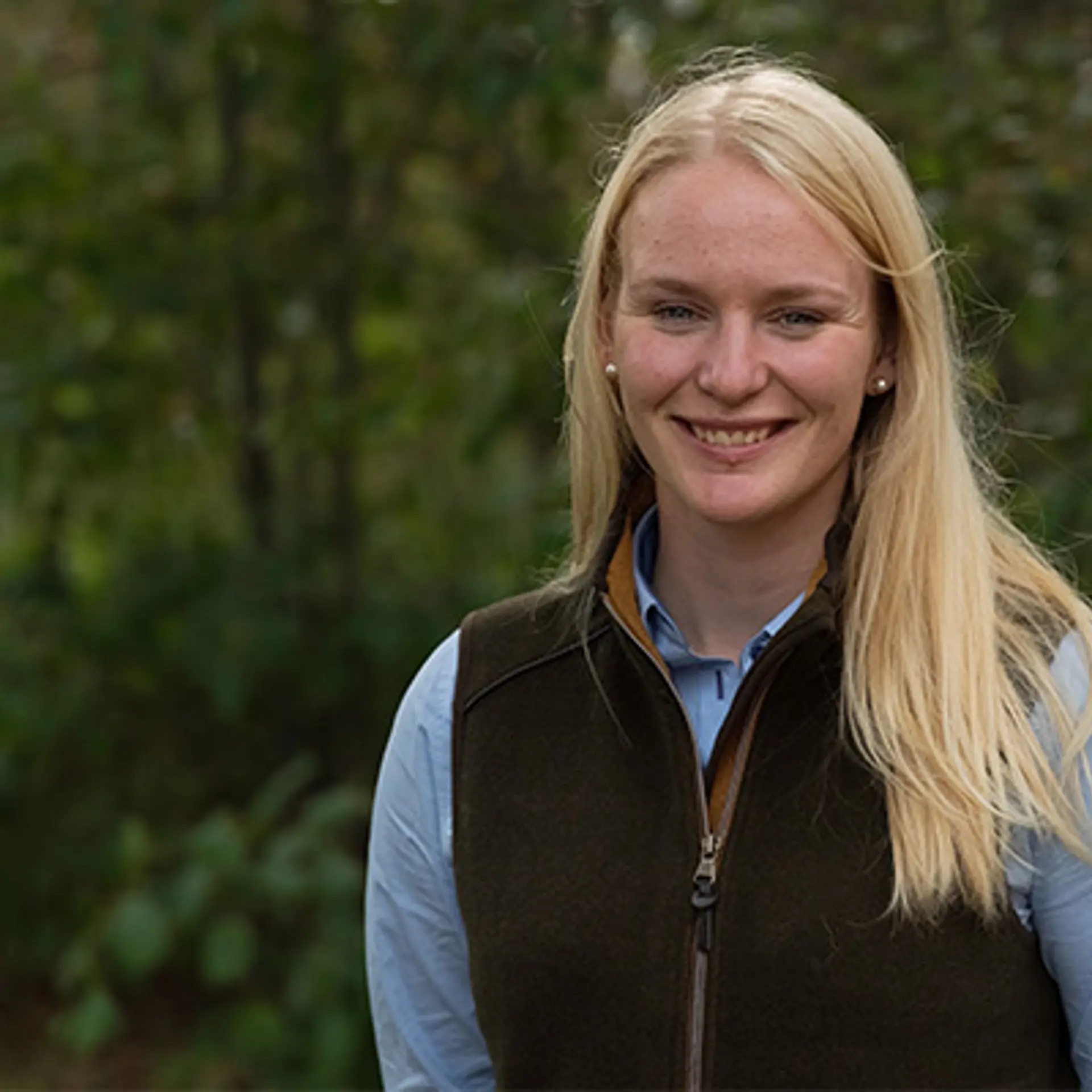 En yngre kvinna med ljust långt hår, blå skjorta och mörkgrön väst ler mot kameran. Hon befinner sig utomhus.