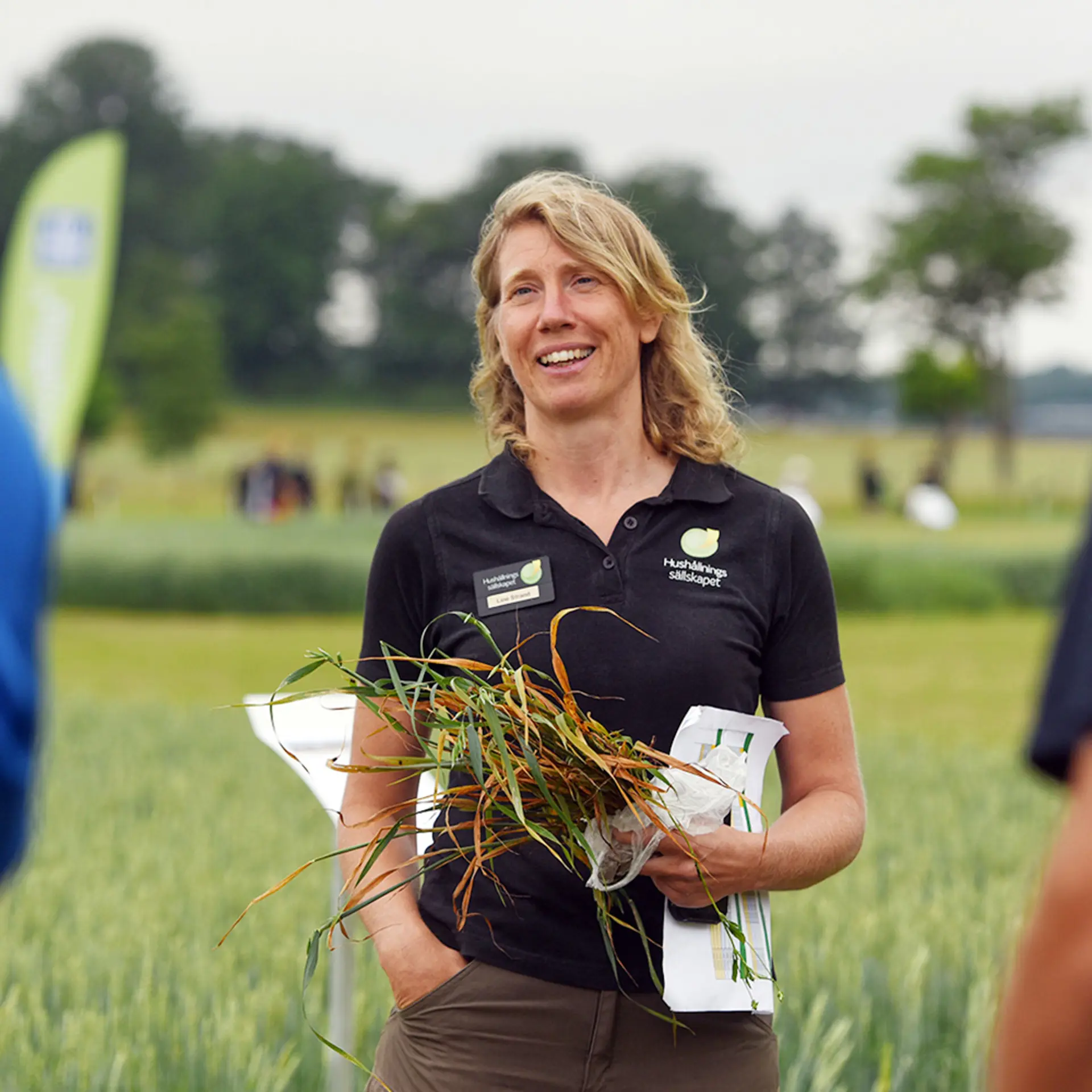 En kvinna med ljust, långt hår står på ett fält och berättar om något. Hon håller en gröda i handen. 