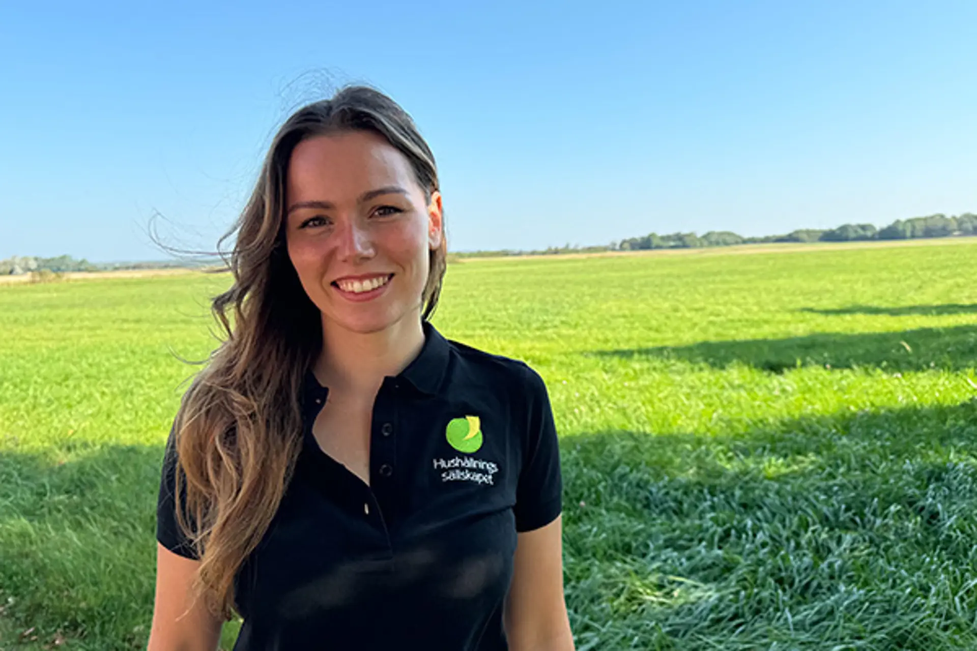 En kvinna i mörkt långt hår och mörk tröja. Ler mot kameran i solen. 